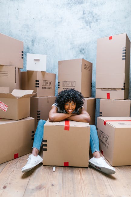 A young woman with curly black hair, wearing a light-colored top and blue jeans, is sitting on a wooden floor inside a room surrounded by numerous cardboard boxes of various sizes. Some boxes have printed labels and red packing tape, indicating they are prepared for moving or packing. She is smiling and resting her chin on a box in front of her, with her arms draped over it, appearing happy and relaxed during a home relocation process. The walls in the background are painted in a textured light blue, and the room is well-lit with natural or artificial light. The scene depicts the packing and moving stage of house removals, highlighting the use of cardboard boxes as part of professional furniture transport and packing services offered by [COMPANY_NAME].