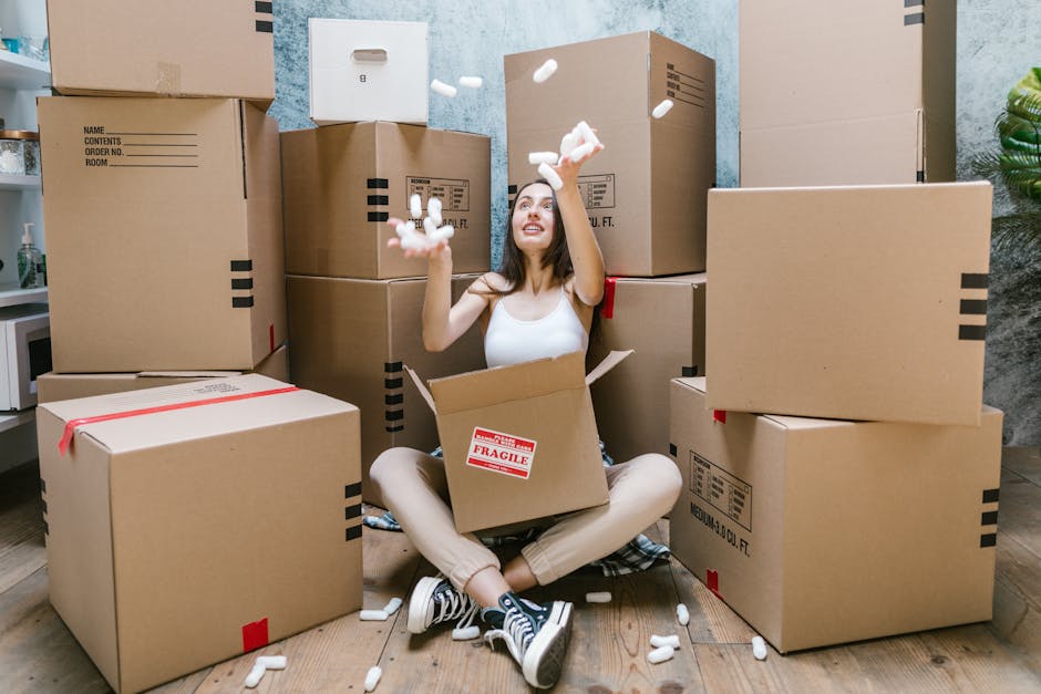 A young woman sitting cross-legged on a wooden floor inside a home surrounded by several large cardboard moving boxes, some sealed with red tape and labeled with contents. She is wearing casual clothing, including a white tank top and sneakers, and is smiling as she throws packing foam peanuts into the air. The boxes are stacked around her, with one open directly in front of her showing additional packing materials inside. The background includes a blue textured wall, a potted plant, and part of a countertop with a microwave, indicating a domestic setting. This scene captures the packing phase of a home relocation process, with visible materials such as cardboard boxes, packing foam, and protective wrapping used for furniture transport, illustrating the logistics involved in house removals often handled by [COMPANY_NAME].