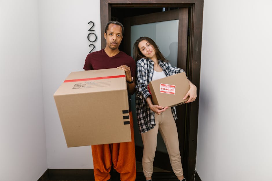 A young woman with curly black hair, wearing a light-colored top and blue jeans, is sitting on a wooden floor inside a room surrounded by numerous cardboard boxes of various sizes. Some boxes have printed labels and red packing tape, indicating they are prepared for moving or packing. She is smiling and resting her chin on a box in front of her, with her arms draped over it, appearing happy and relaxed during a home relocation process. The walls in the background are painted in a textured light blue, and the room is well-lit with natural or artificial light. The scene depicts the packing and moving stage of house removals, highlighting the use of cardboard boxes as part of professional furniture transport and packing services offered by [COMPANY_NAME].