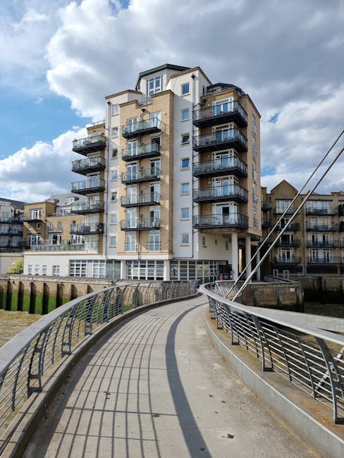 A multi-storey residential building located on a riverside in Kingston upon Thames, featuring multiple balconies with glass railings and cream-colored external walls. The building is positioned behind a curved, paved pathway with metal railings along the edge, which runs parallel to the river. The sky above is partly cloudy with patches of blue. The scene captures the urban environment typical of house removals or property relocations, with the building serving as a backdrop for potential furniture transport or packing and moving activities facilitated by house removals specialists such as manwithvankingstonuponthames.co.uk.