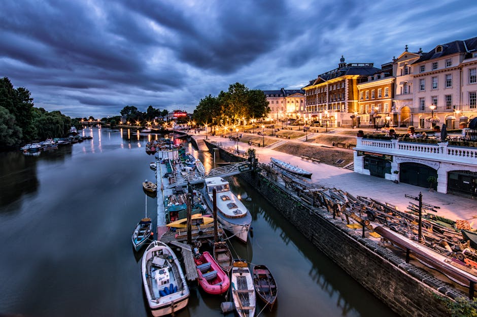 A multi-storey modern residential building with curved balconies and large glass windows, situated along the riverside in Kingston upon Thames. The building's facade features yellow brickwork and dark metal railings on the balconies. In the foreground, several boats, including a white vessel and a blue and white boat named 'Leonid,' are docked on the riverbank. The surrounding area includes lush green trees and an outdoor seating area with tables and umbrellas, indicating a riverside setting. Bright daylight and a partly cloudy sky illuminate the scene, which exemplifies a typical riverside residential environment suitable for house removals and furniture transport services provided by companies like manwithvankingstonuponthames.co.uk. The scene captures the essence of a well-maintained urban riverside with ongoing activities related to home relocation and moving logistics, reflecting the picturesque setting for a Kingston riverside house move plan.