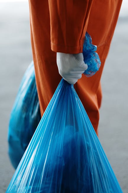 A person wearing orange overalls and a white glove is holding a blue plastic rubbish bag filled with waste. The individual is standing outdoors on a paved surface with a grey background, possibly a driveway or loading area. Nearby, there are additional rubbish bags and cardboard boxes, some wrapped in plastic, indicating a cleanup or moving process. The scene suggests preparations for a home relocation or household waste collection, with the focus on the removal of unwanted items. The person’s grip on the bag is firm, and the items appear ready to be transported, reflecting typical activities associated with furniture transport, packing, and moving logistics. The natural daylight illuminates the scene clearly, highlighting the materials and objects involved in the waste or removal process, which aligns with the context of Kingston Council rules for skips and bulky waste removals, managed by [COMPANY_NAME].