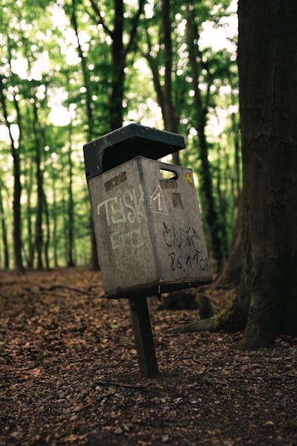 A grey metal postal box mounted on a tilted black metal post, situated on a dirt path within a wooded area with tall trees and dense green foliage in the background. The postal box features a slanted black lid, a rectangular slot for letters, and graffiti markings on its surface. The ground is covered with soil and small rocks, with some leaves scattered around. Natural light filters through the tree canopy, illuminating the scene. This image illustrates outdoor mail collection in a natural environment, relevant to moving or home relocation contexts like those mentioned on the Kingston Council rules for skips and bulky waste removals page at manwithvankingstonuponthames.co.uk, where proper waste and package handling are part of the logistics process.