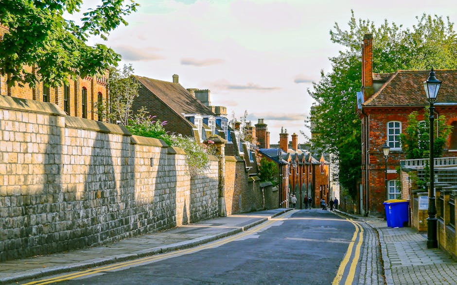 A quiet residential street near Kingston station during daylight, featuring a sloped asphalt road with double yellow lines on the left side, a stone pavement sidewalk on the right, and a stone wall lining the left side. Several brick and stone houses with chimneys, dormer windows, and sloped roofs are visible, with some having small front gardens or terraces. A traditional black street lamp is situated on the right sidewalk, near a blue wheelie bin. Tall leafy trees provide greenery along the street, and a few pedestrians are seen walking in the distance. The scene is well-lit with natural light, highlighting the textures of the bricks, stones, and foliage, illustrating a typical quaint, historic neighbourhood supporting home relocations and moving logistics by providing a clear view of the environment for furniture transport and packing preparations, as referenced in the Kingston station removals guide.