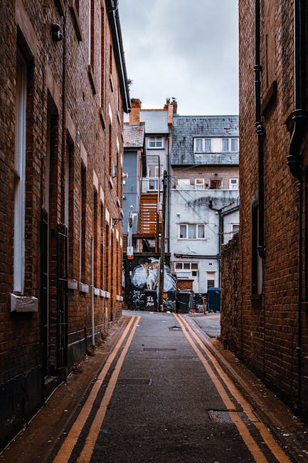 A narrow alleyway between two brick buildings in Kingston upon Thames, showing a loading area for house removals. The alley features rain-slicked pavement marked with double yellow lines along the edges. In the background, a moving van is partially visible with its rear door open, and several cardboard boxes, plastic-wrapped furniture, and packing materials are placed on the pavement near the van. The surrounding buildings include residential structures with windows, and a small wooden staircase leads to an upper-floor door of one building. Overhead, the sky is cloudy, indicating overcast weather, with professional furniture blankets and moving equipment such as trolleys and straps possibly present, supporting a home relocation process. This setup reflects a typical scene during a furniture transport and packing and moving operation in a city environment, as handled by companies like [COMPANY_NAME].