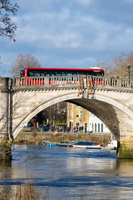 A red and white double-decker bus is traveling across a stone arch bridge over the River Thames in Kingston upon Thames. The bridge features decorative stone balustrades with a classical design. Below, the river reflects the blue sky and scattered clouds, with small boats docked along the shoreline on the opposite bank. In the background, there are residential buildings and trees with bare branches, suggesting late autumn or early winter. The scene captures a typical daytime view of a home relocation area, with the infrastructure supporting furniture transport and moving logistics within the town. The image is well-lit with natural sunlight, emphasizing the clarity of the bridge, river, and surrounding environment, illustrating the urban setting integral to organized house removals and packing and moving operations in Kingston upon Thames.