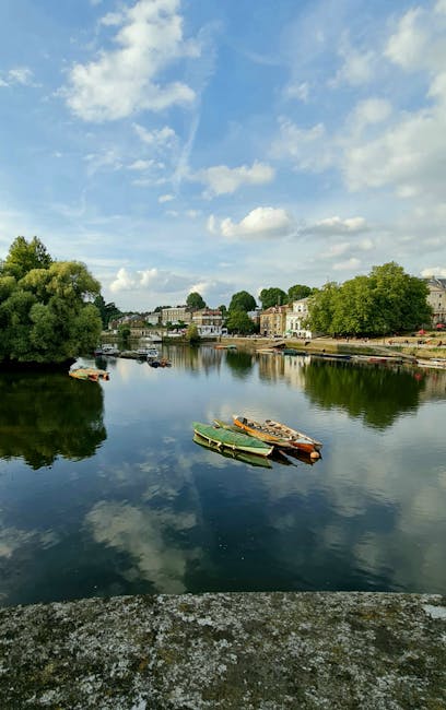 A panoramic view of the River Thames in Kingston upon Thames during daytime, featuring calm water reflecting a partly cloudy sky with scattered white clouds and blue patches. On the river, several small boats with green, orange, and wooden surfaces are moored close to the bank. Along the riverbank, there are rows of residential buildings and trees, with some greenery extending near the water's edge. The scene captures a peaceful, outdoor environment suitable for relaxed viewing or river transport activities. The image's natural lighting highlights the vibrant colors of the boats and the lush foliage on the surrounding trees, with a subtle focus on the serenity of the riverside setting, which could relate to home relocation or transportation services provided by [COMPANY_NAME] in Kingston upon Thames.