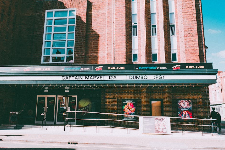 A view of the exterior of the Rose Theatre in Kingston upon Thames, showing the building's brick facade with a large, rectangular window on the upper level. The theatre's marquee displays information about upcoming shows, including the musical 'Blood Brothers' from May 10 to June 2, and the film 'Dumbo' rated PG, with the title 'Captain Marvel 12A' also visible. In front of the theatre, there is a pavement with a white metal railing leading up to the entrance, which consists of double glass doors. To the right side of the entrance, a person is walking along the sidewalk, and advertisements or posters are attached to the brick wall near the door. The lighting suggests a bright, clear day, ideal for home relocation or furniture transport activities as often coordinated by professional removals teams like [COMPANY_NAME]. This scene may be used in the context of planning or carrying out house removals, packing, and moving logistics to or from Kingston upon Thames, emphasizing the importance of careful planning during the loading process of furniture and boxes onto transport vehicles.