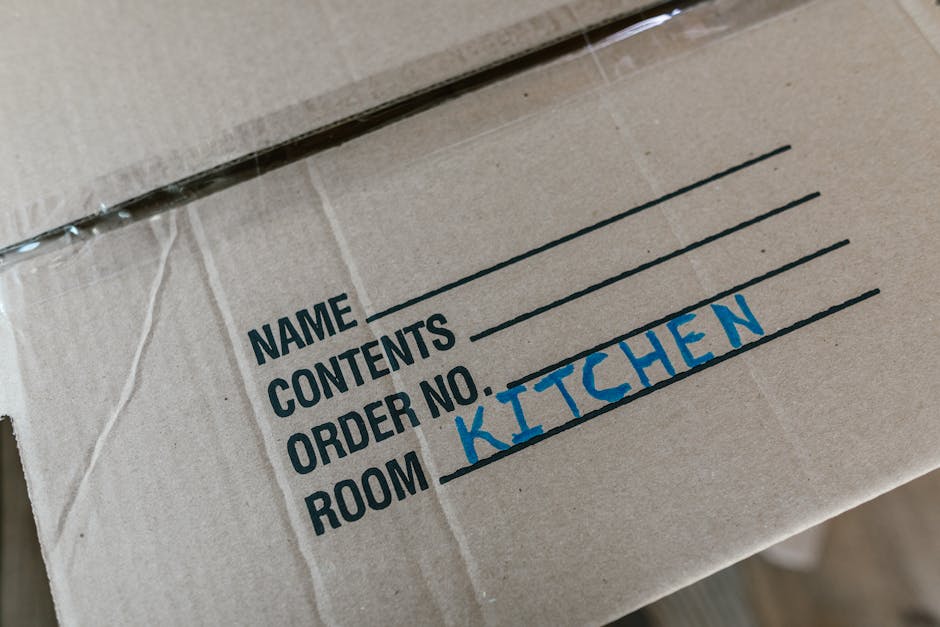 Close-up view of a cardboard moving box with a white label on top, displaying sections for 'Name', 'Contents', 'Order No.', and 'Room', with the word 'KITCHEN' handwritten in blue marker in the 'Room' section. The box appears to be part of a house removal or relocation process, likely inside a home or in the process of being packed, with other boxes and packing materials not visible in this frame. The lighting is neutral, highlighting the label and the plain cardboard surface, which is consistent with professional packing and furniture transport activities as seen in house moving and removals services, such as those by manwithvankingstonuponthames.co.uk.