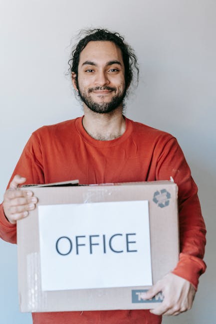 A man with dark, curly hair and a beard is standing indoors against a plain light-colored wall, smiling and holding a cardboard box labeled 'OFFICE' with both hands. The box appears to contain office supplies or equipment and is secured with clear packing tape. He is wearing a red long-sleeved shirt. In the background, part of a doorway or hallway is visible, suggesting the setting is at the entrance or interior of an office or home environment. This image reflects the process of office furniture or equipment packing as part of a house or business relocation, with clean, well-organized packaging materials. The natural lighting highlights the subject and the box clearly, supporting the theme of furniture transport, packing, and moving logistics, which aligns with professional removal services such as those offered by [COMPANY_NAME].